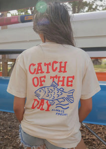 tan tee shirt with "catch of the day" fish graphic printed on back and "a real catch" printed centered on front 
