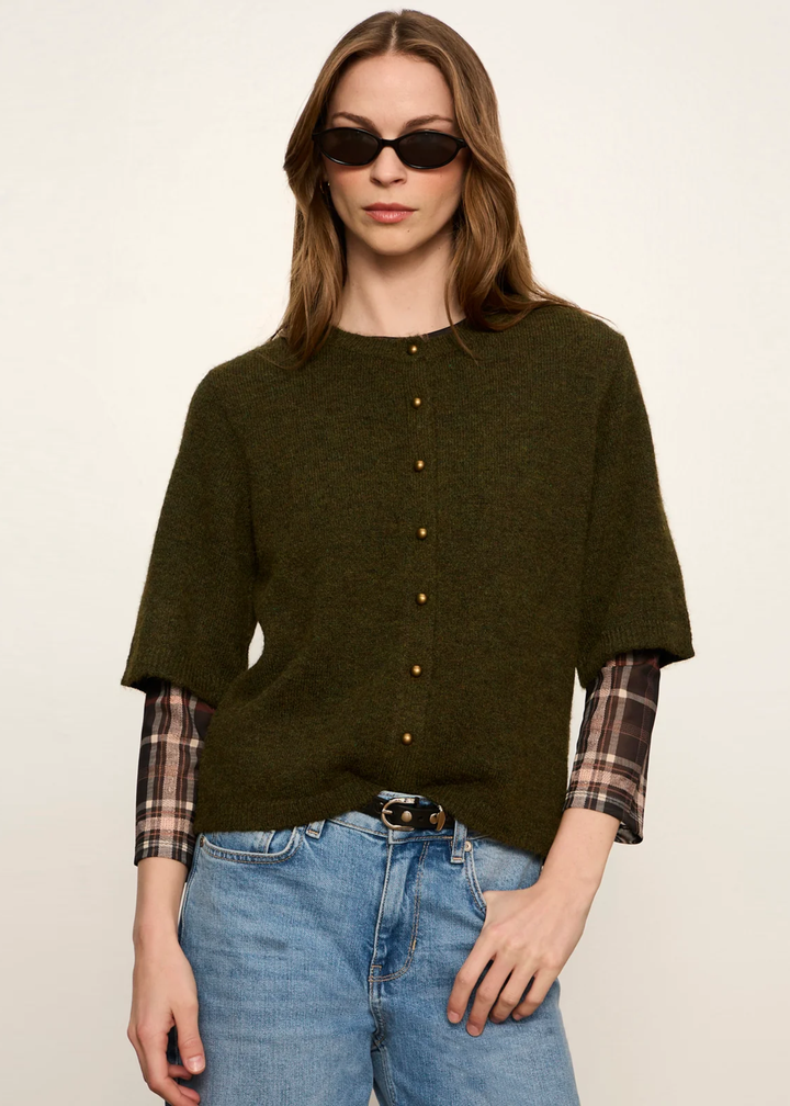 A woman modeling an olive green short-sleeve knit top with a button-down front, layered over a plaid shirt, paired with blue jeans and sunglasses, against a plain off-white background.