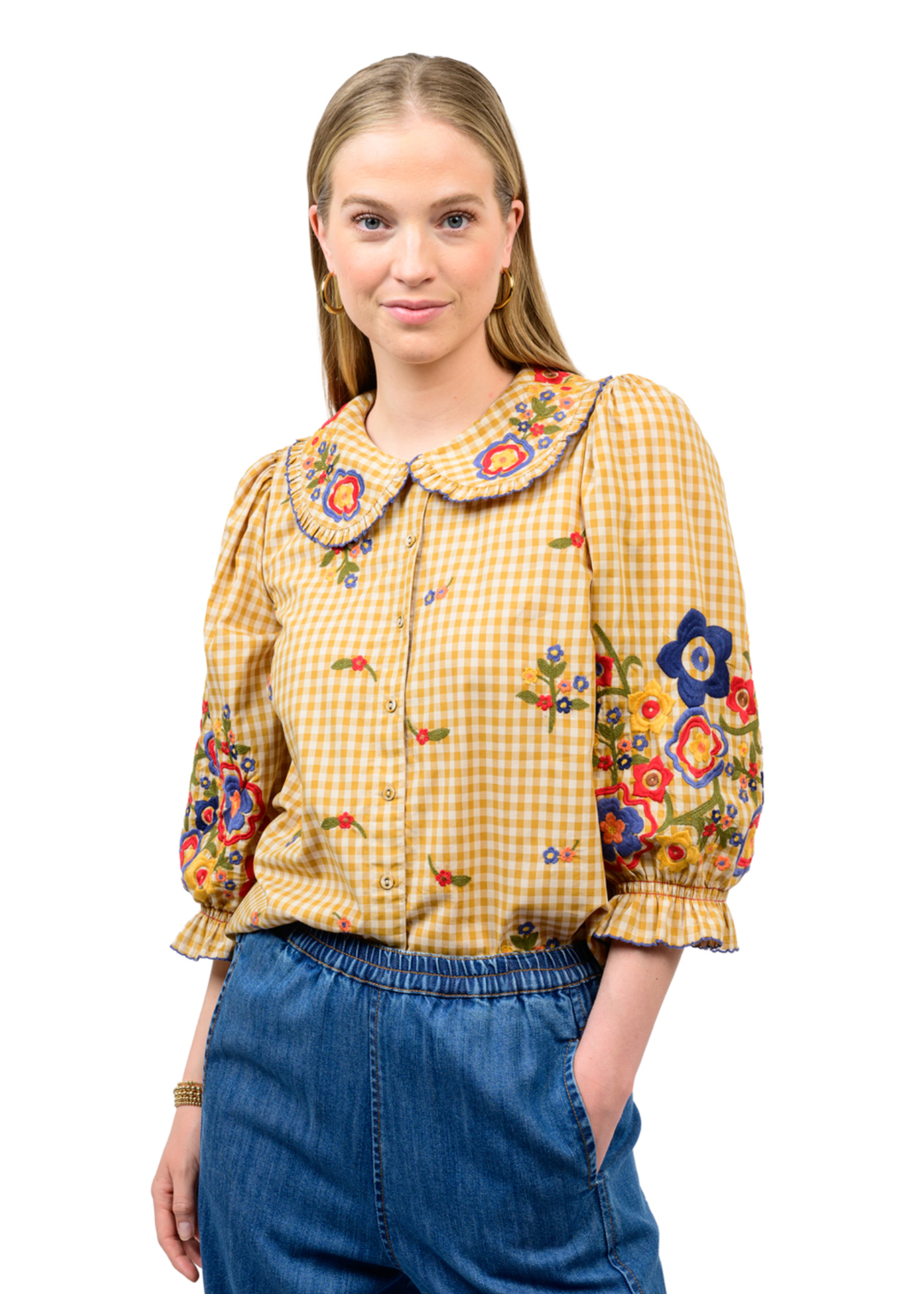 A woman modeling a yellow and white gingham button-up blouse with colorful floral embroidery on the collar, cuffs, and front, paired with blue jeans against a plain white background.