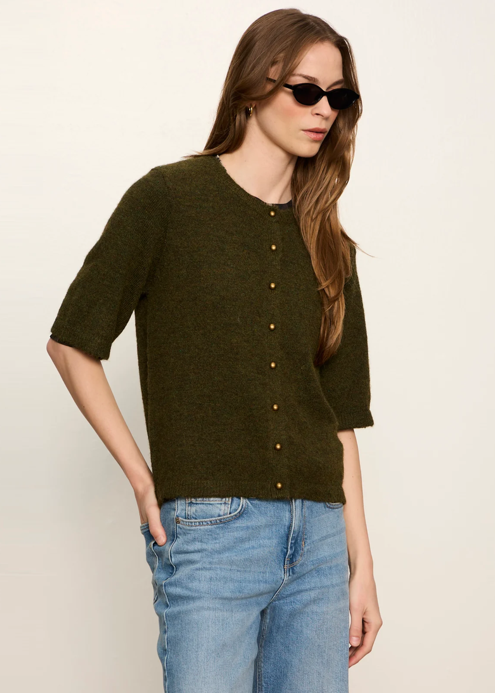 A woman modeling a short-sleeve olive green knit top with a button-down front, paired with light blue jeans, against a plain off-white background.