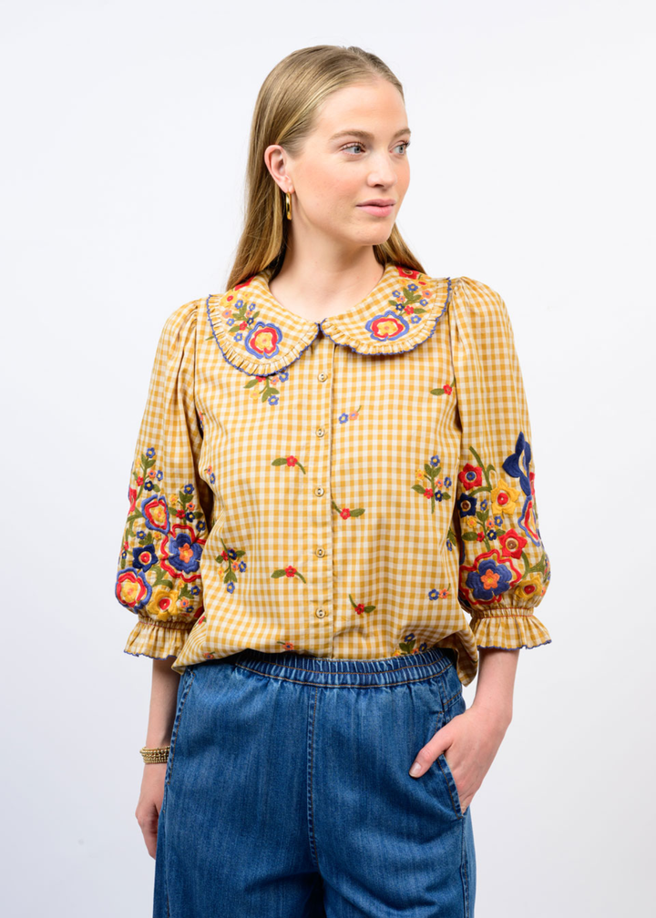 A woman modeling a yellow and white gingham blouse with colorful floral embroidery on the collar, cuffs, and body, paired with blue jeans against a plain white background.