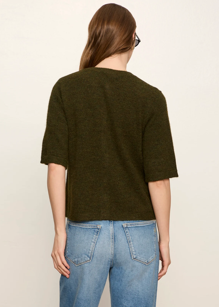 A woman viewed from the back wearing a dark olive green short-sleeve knit top and light blue jeans against a plain off-white background.