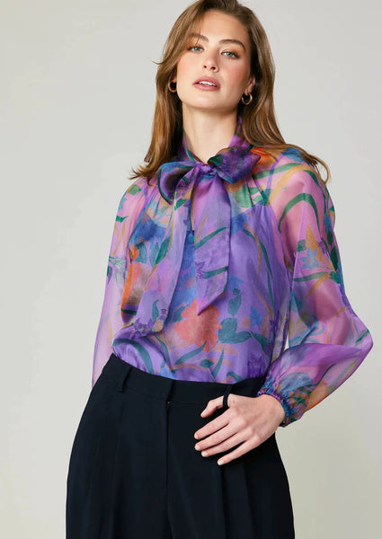 Sheer purple floral blouse with dramatic neck bow and puff sleeves layered over a simple built in dark purple cami 