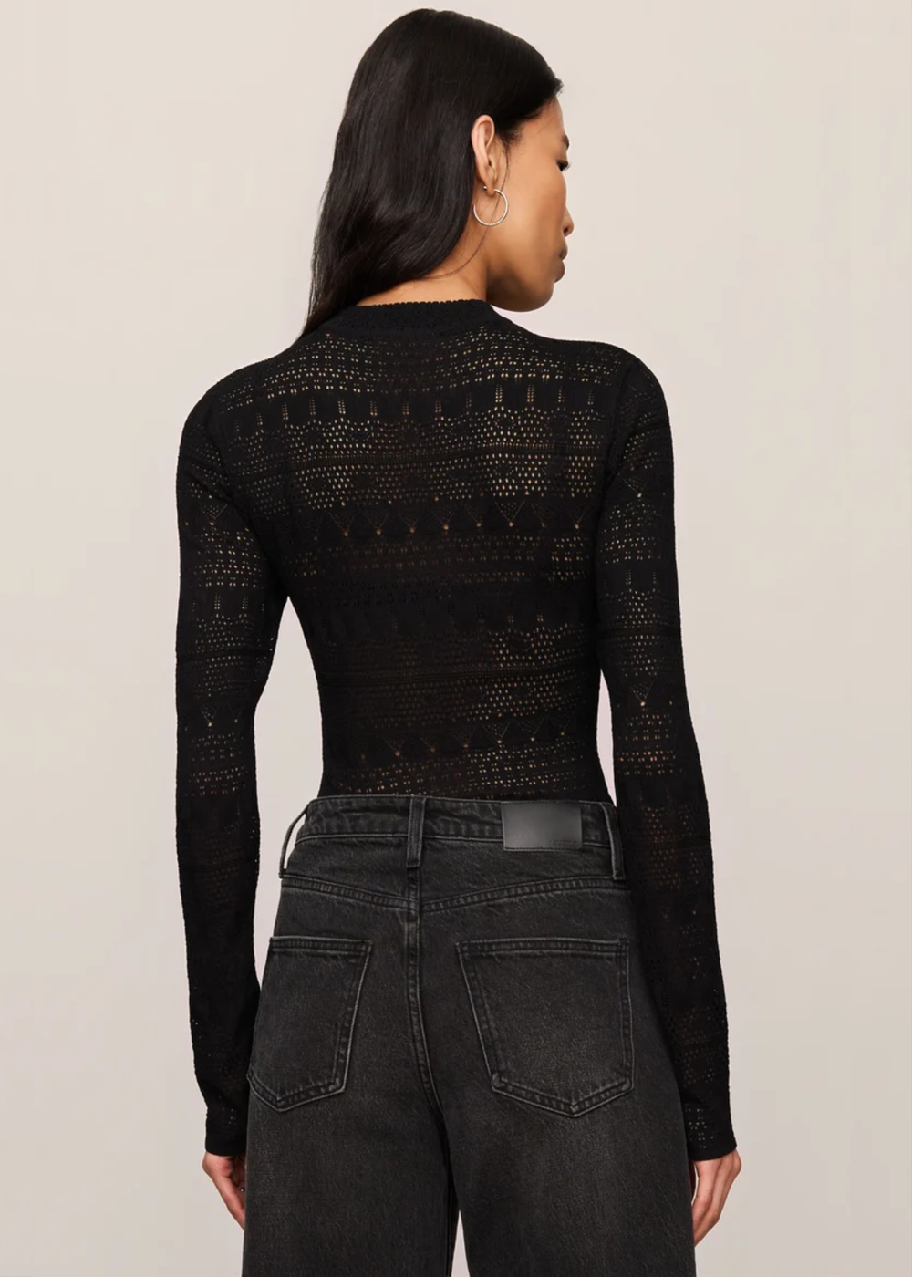 A woman shown from the back wearing a black long-sleeve knit top with a textured openwork pattern, paired with dark gray jeans, against a plain light beige studio background.
