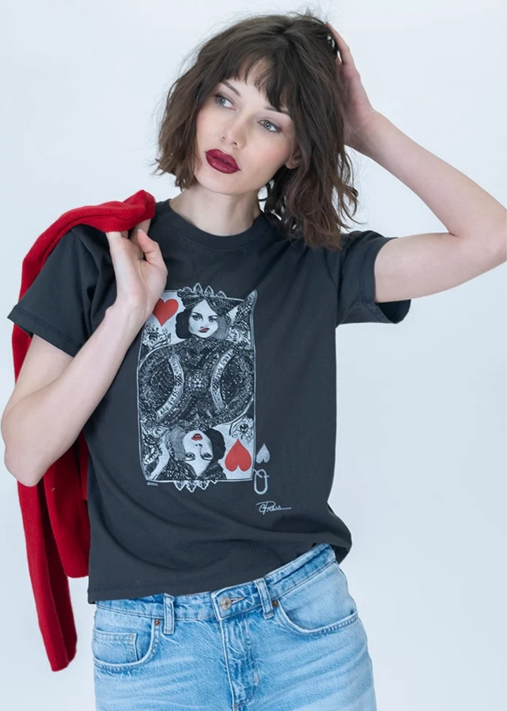 A woman modeling a black graphic t-shirt featuring a Queen of Hearts playing card design, paired with light blue jeans and holding a red jacket over her shoulder against a plain white background.