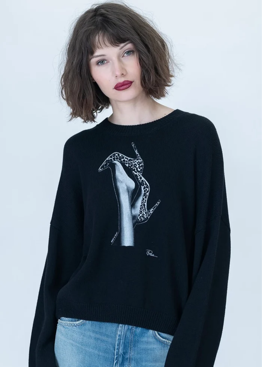 A woman with short brown hair and dark red lipstick wears a black crewneck sweater featuring a white and gray graphic of a hand holding a penguin, paired with light blue jeans, against a light gray background.
