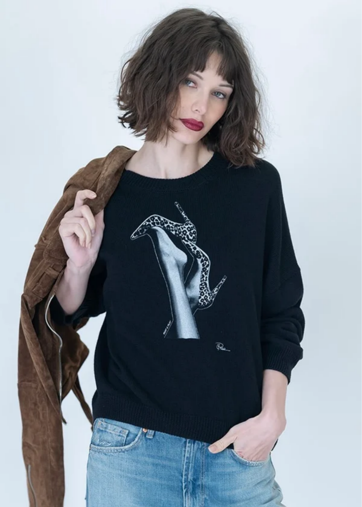 A woman wearing a black crewneck sweater with a graphic print of a high-heeled shoe, styled with light blue jeans and a brown suede jacket draped over her shoulder, against a plain light gray background.