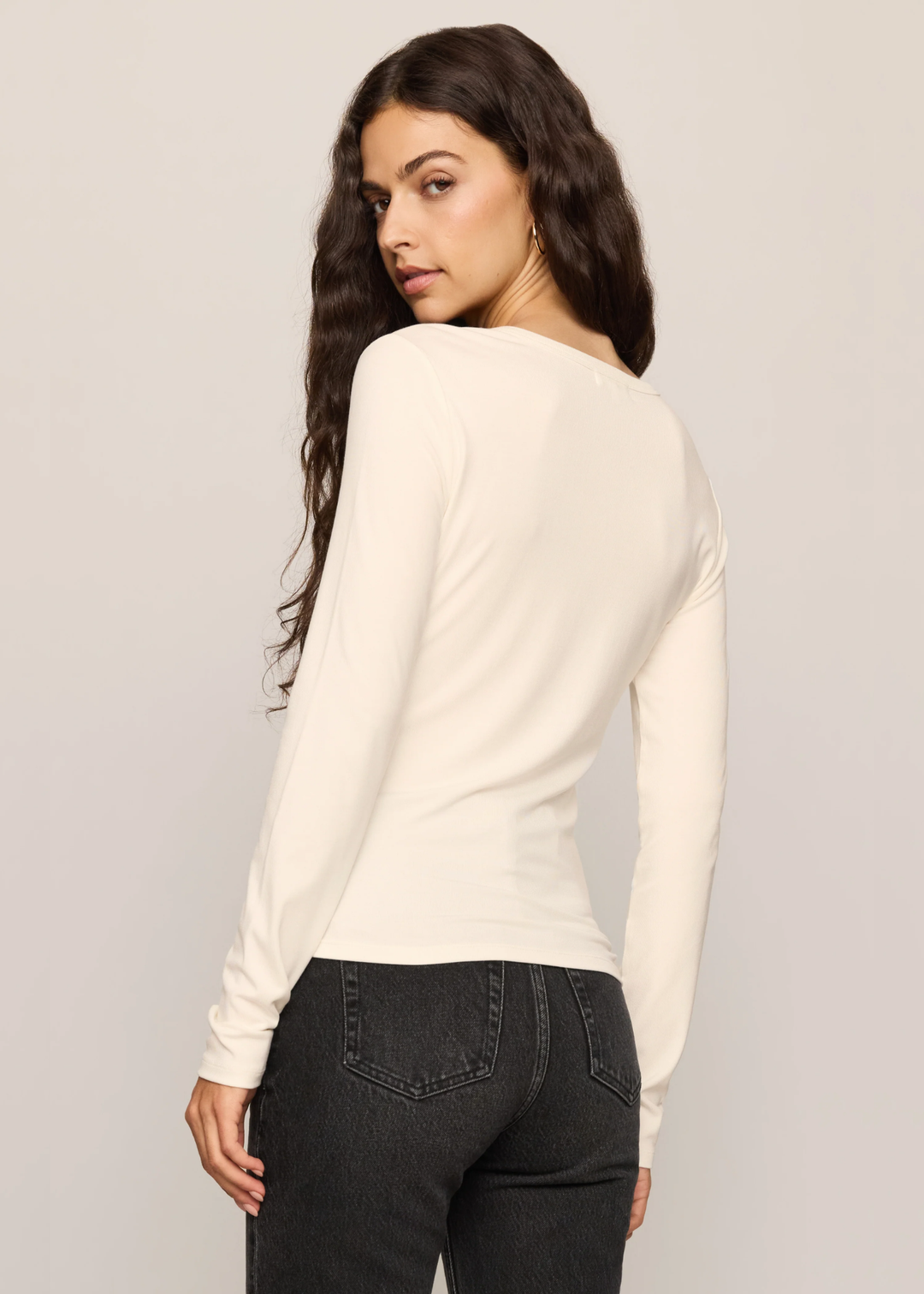 A woman with long dark hair is shown from the back wearing a fitted off-white long-sleeve top and dark gray jeans against a light gray studio background.