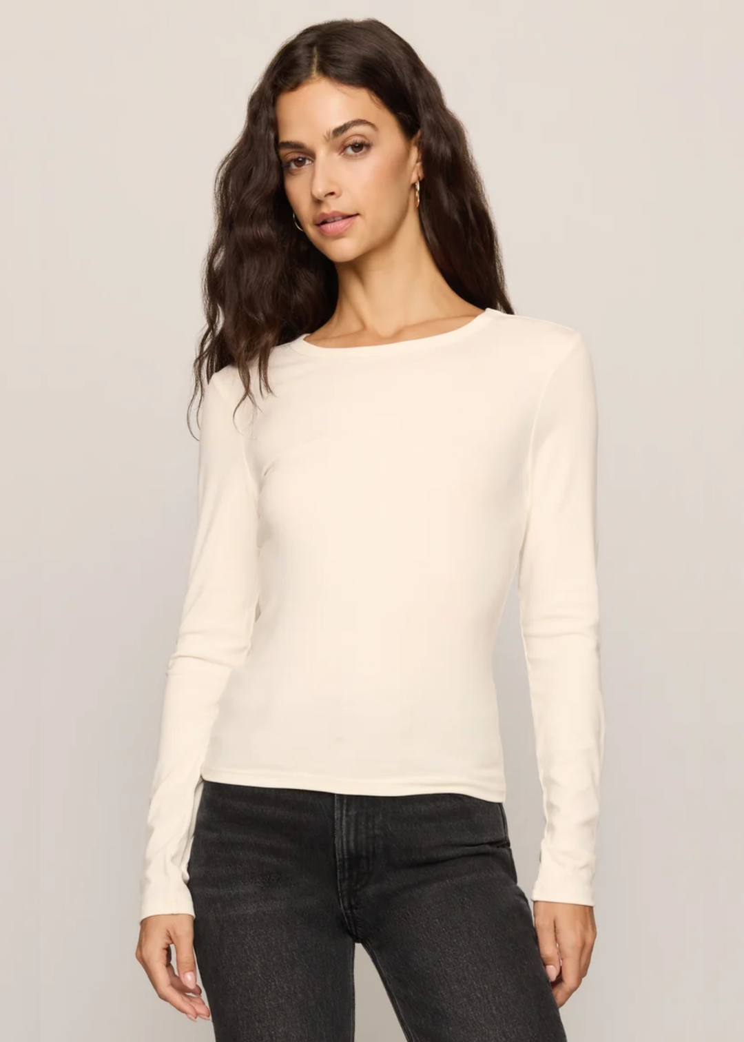 A woman modeling a fitted, long-sleeve crewneck top in off-white, paired with dark gray jeans, against a plain light gray studio background.