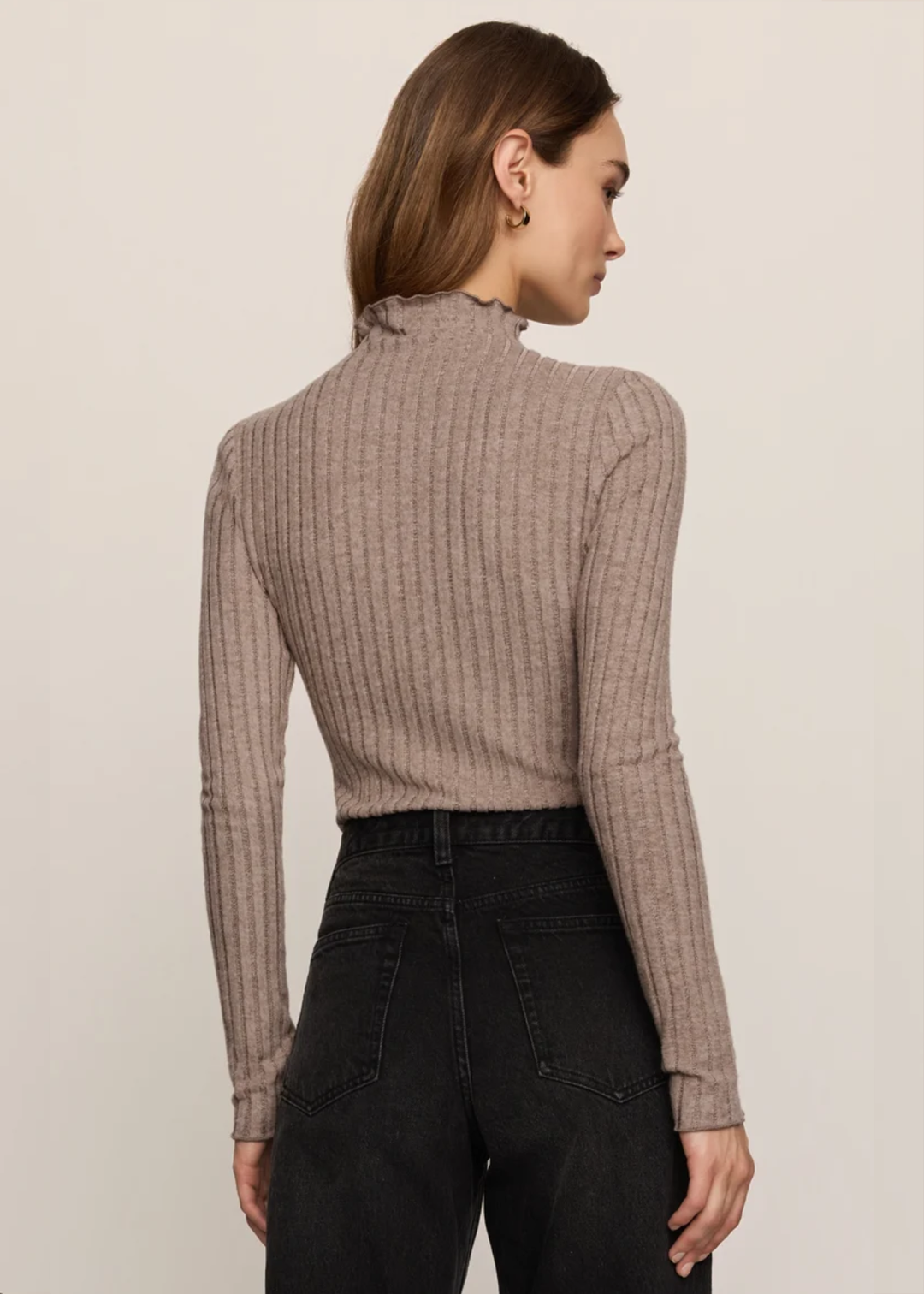 A woman shown from the back wearing a ribbed long-sleeve mock neck top in taupe and black jeans, against a plain light beige studio background.