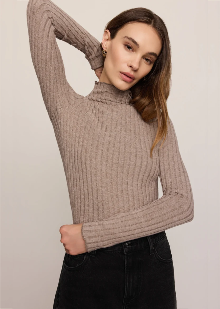 A woman wearing a ribbed knit mock neck sweater in taupe, paired with black jeans, posing against a neutral light gray background.