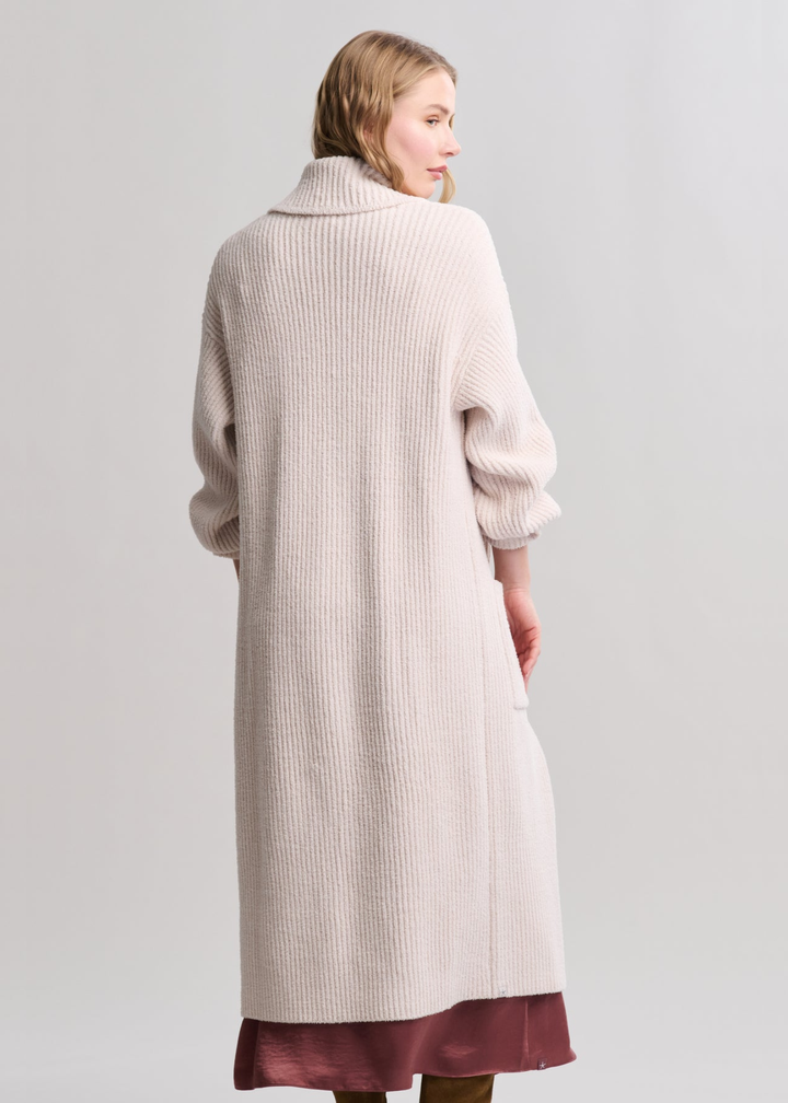 Woman turned around wearing a long cream coat over a mauve maxi dress standing in front of a grey background