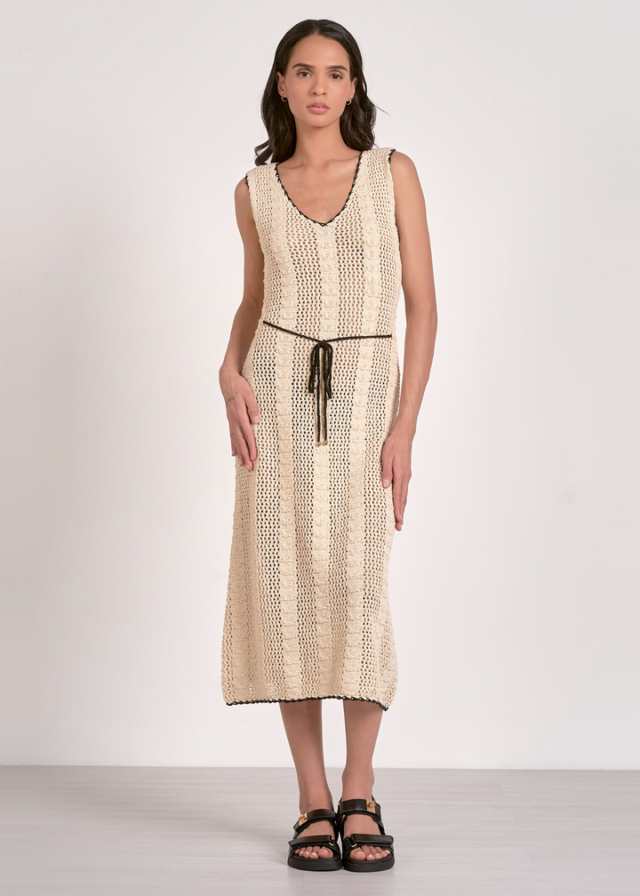 A woman wearing a tan crochet dress with black trim and a black belt in front of a white background