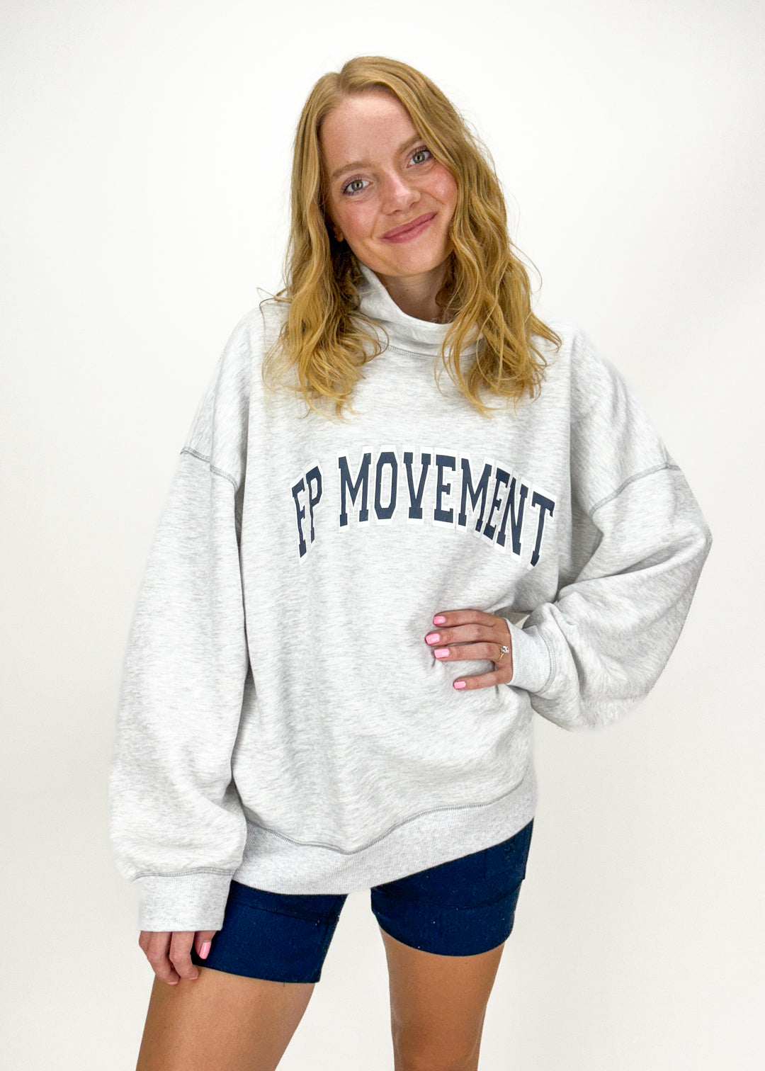 woman wearing a heather grey mock neck sweatshirt with navy blue "FP MOVEMENT" graphic on chest with coordinating navy blue biker shorts