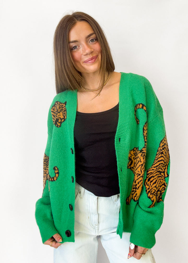 A woman wearing a bright green knit cardigan with large tiger prints on the sleeves and shoulders, styled over a black top and white jeans against a plain white background.