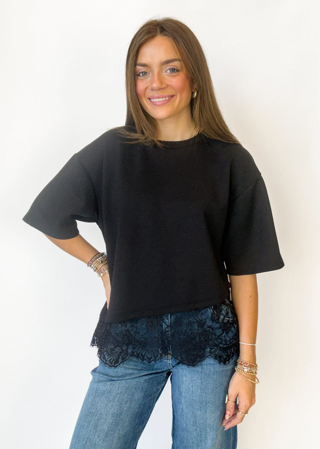 A woman modeling a black oversized short-sleeve t-shirt with a black lace hem, paired with blue jeans, against a plain white background.