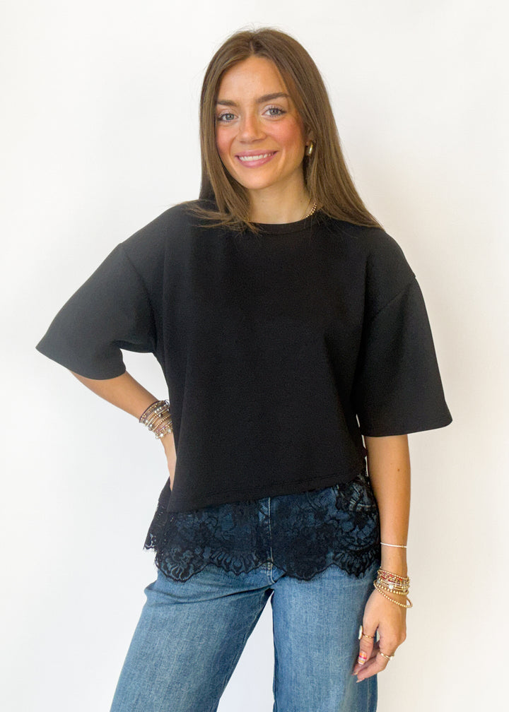 A woman modeling a black oversized short-sleeve t-shirt with a black lace hem, paired with blue jeans, against a plain white background.
