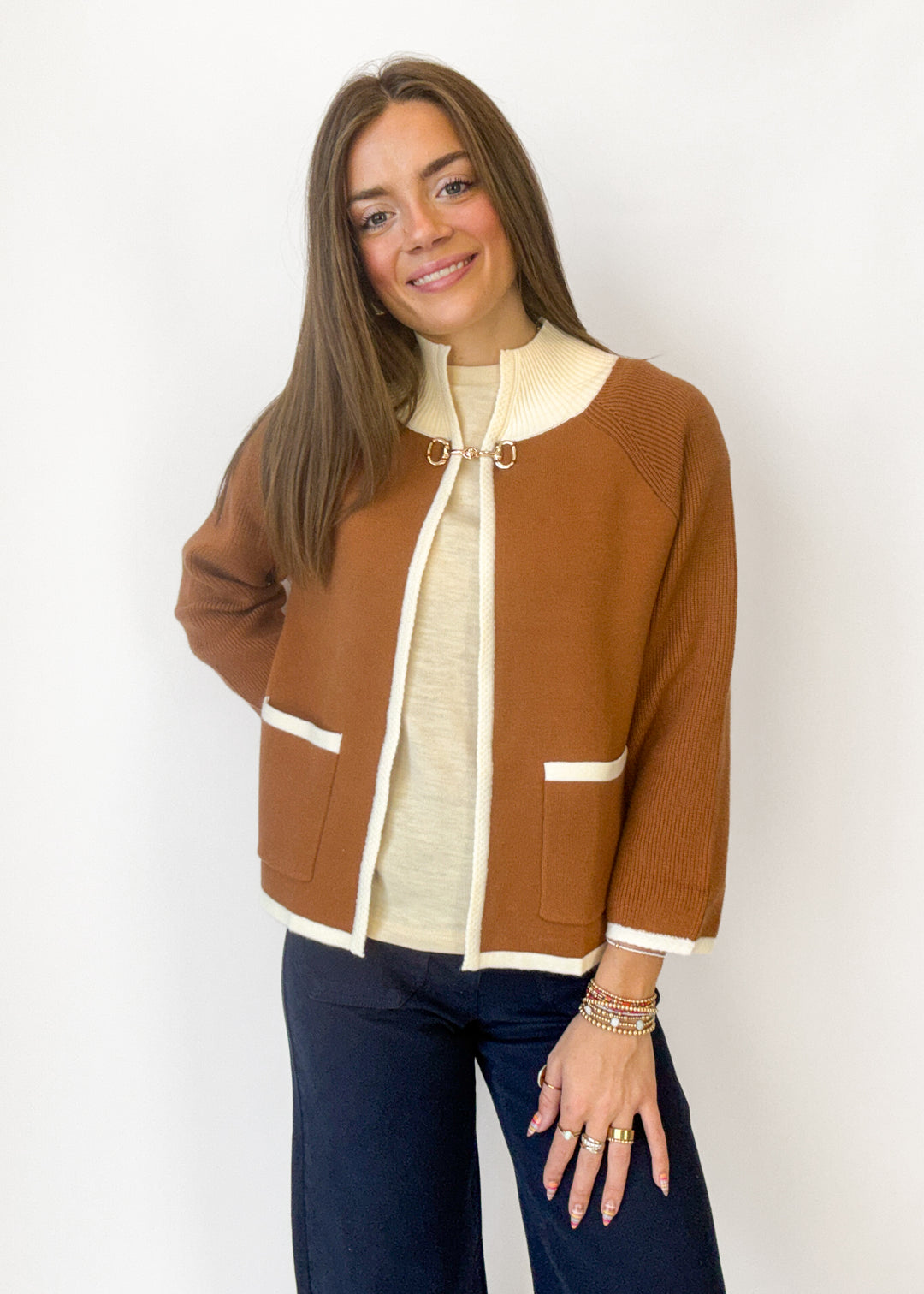 A woman modeling a brown knit zip-up cardigan with cream trim over a cream top and dark blue jeans, standing against a plain white background.