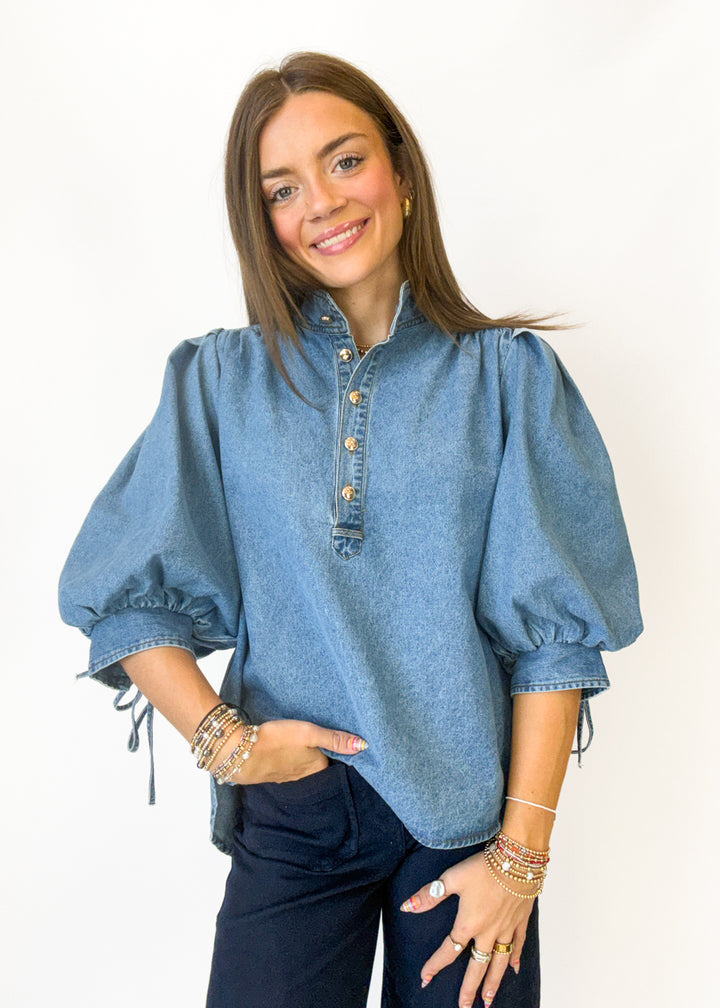 A woman modeling a light blue denim puff-sleeve blouse with a buttoned stand collar and dark navy pants, standing against a plain white background.