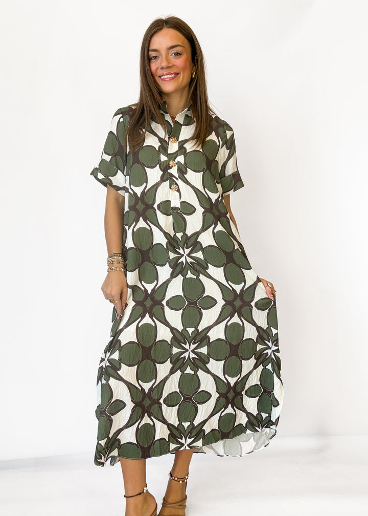 A woman modeling a short-sleeve midi dress with a green and white geometric floral pattern, standing against a plain white studio background.