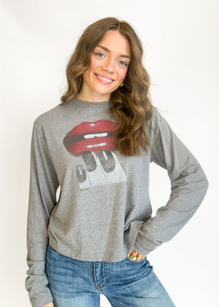 A woman wearing a heather gray long-sleeve crewneck sweatshirt with a graphic of red lips biting a sign that says '99' on the front, paired with blue jeans, against a plain white background.