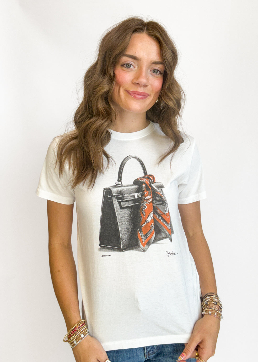 A woman wearing a white graphic t-shirt featuring a black handbag with an orange and black patterned scarf, paired with blue jeans and gold bracelets, against a plain white background.