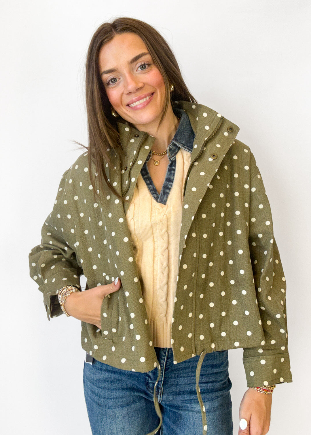 A woman modeling an olive green jacket with white polka dots, worn over a cream cable-knit sweater and blue jeans against a plain white background.
