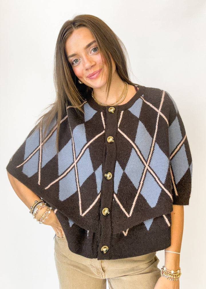A woman modeling a brown and light blue argyle knit poncho cardigan with gold buttons, paired with beige pants, against a plain white background.