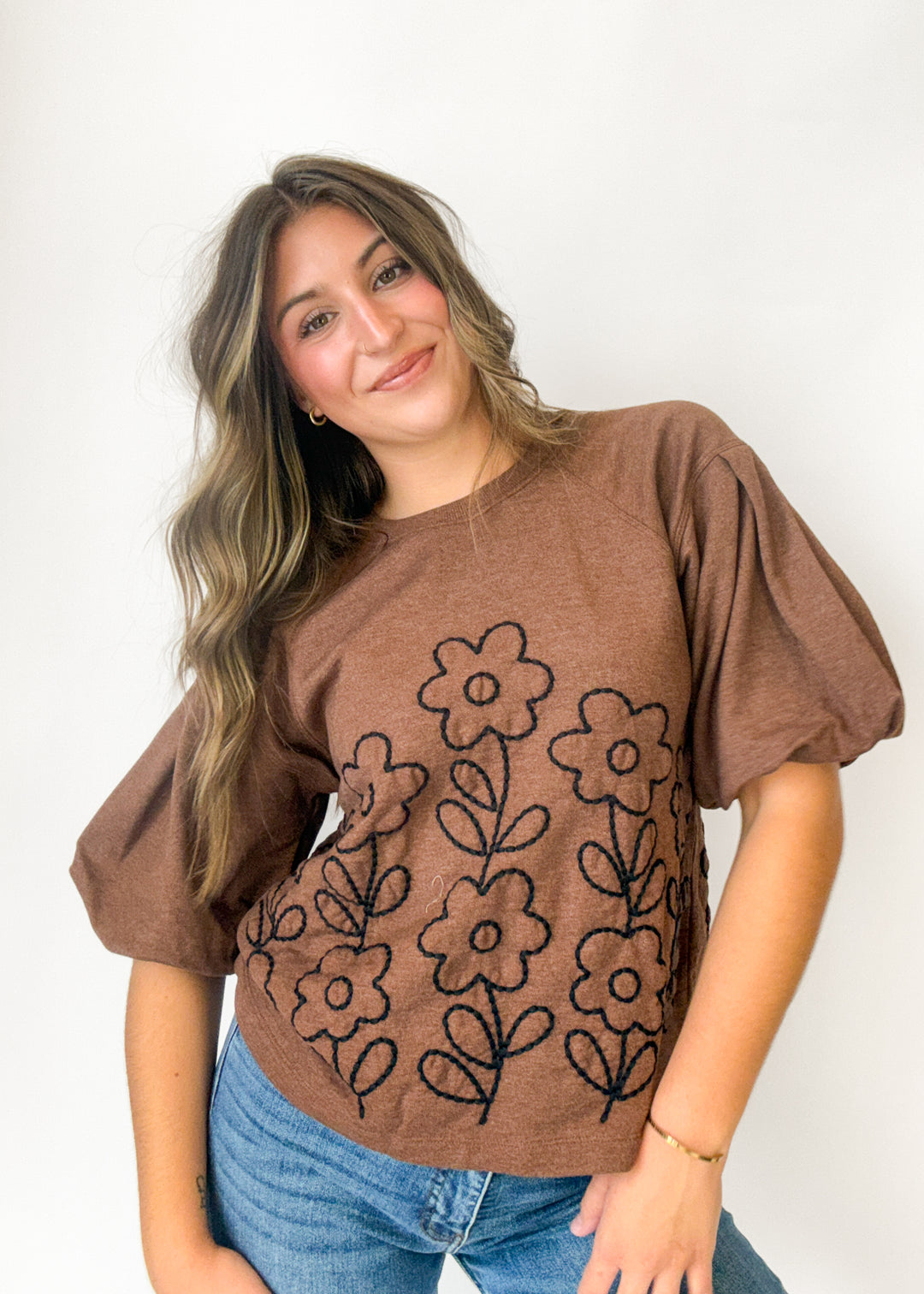 A woman wearing a brown short-sleeve t-shirt with black embroidered flowers, paired with blue jeans, posing against a plain white background.
