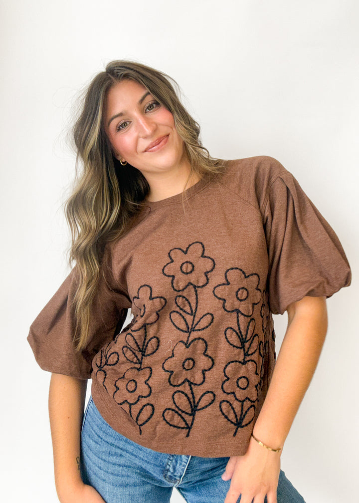 A woman wearing a brown short-sleeve t-shirt with black embroidered flowers, paired with blue jeans, posing against a plain white background.