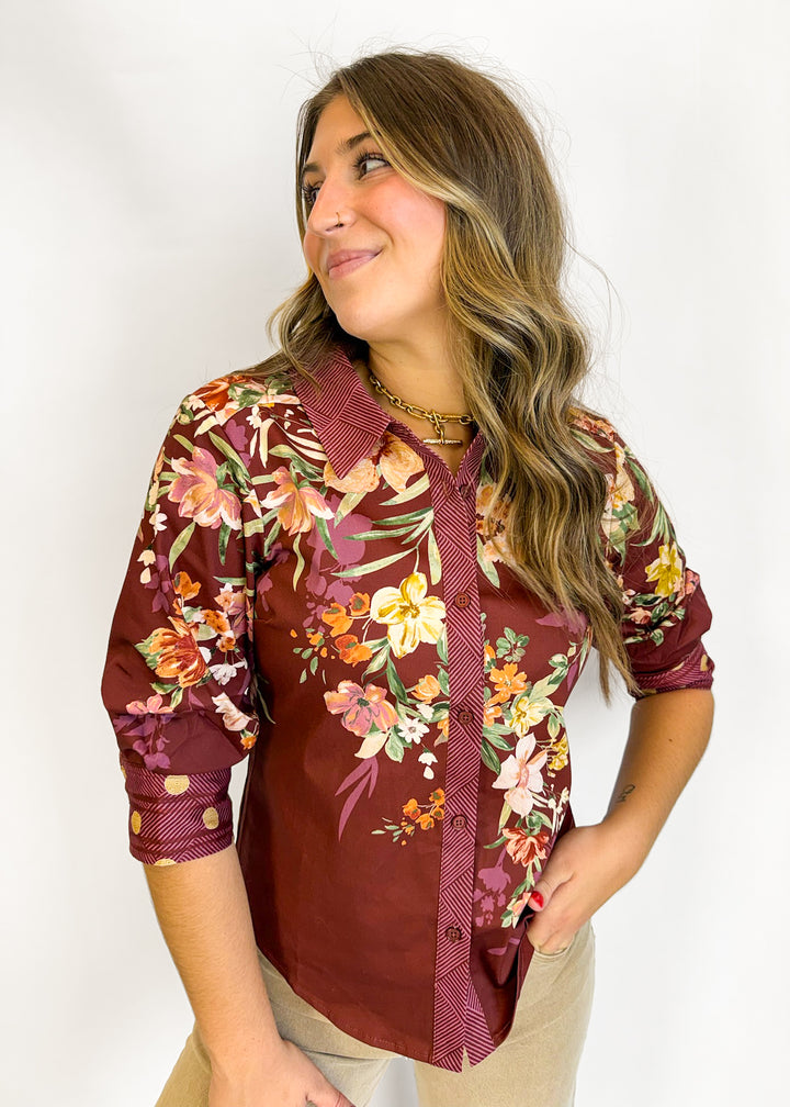 A woman wearing a burgundy button-up shirt with a vibrant multicolor floral print, paired with beige pants, photographed against a plain white studio background.