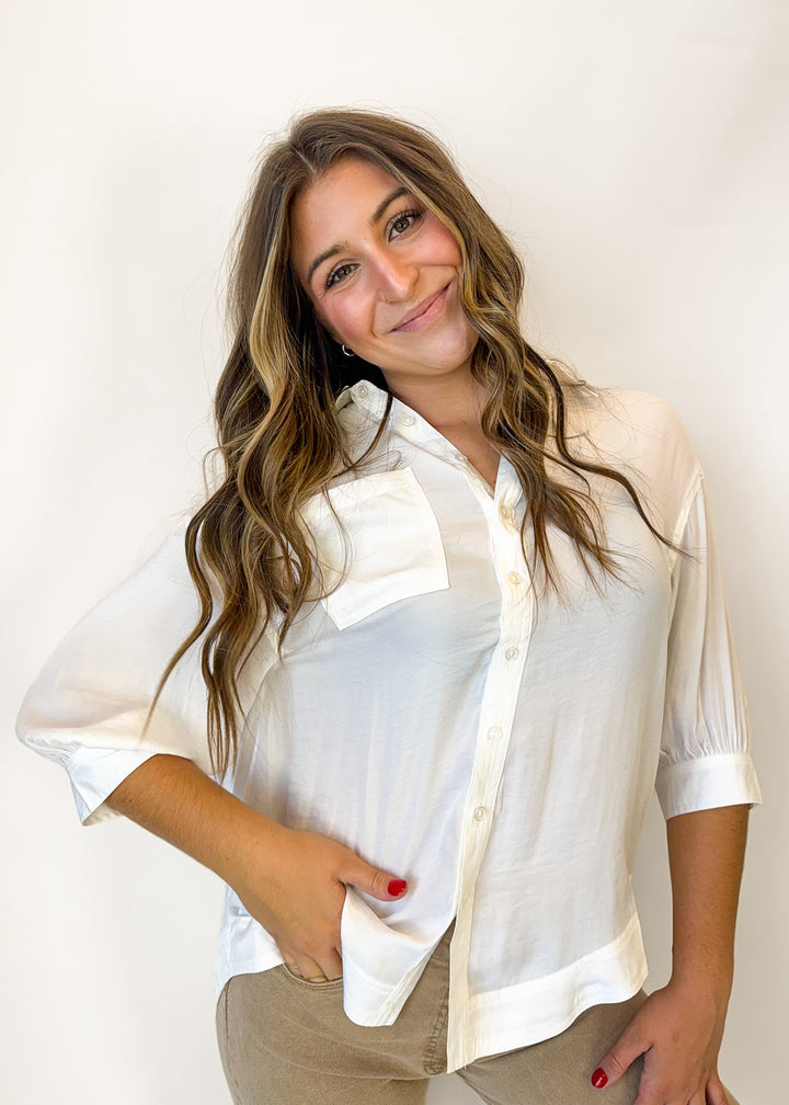 A woman with long wavy brown hair wearing a white semi-sheer three-quarter sleeve button-up shirt and beige pants, posing against a plain white studio background.