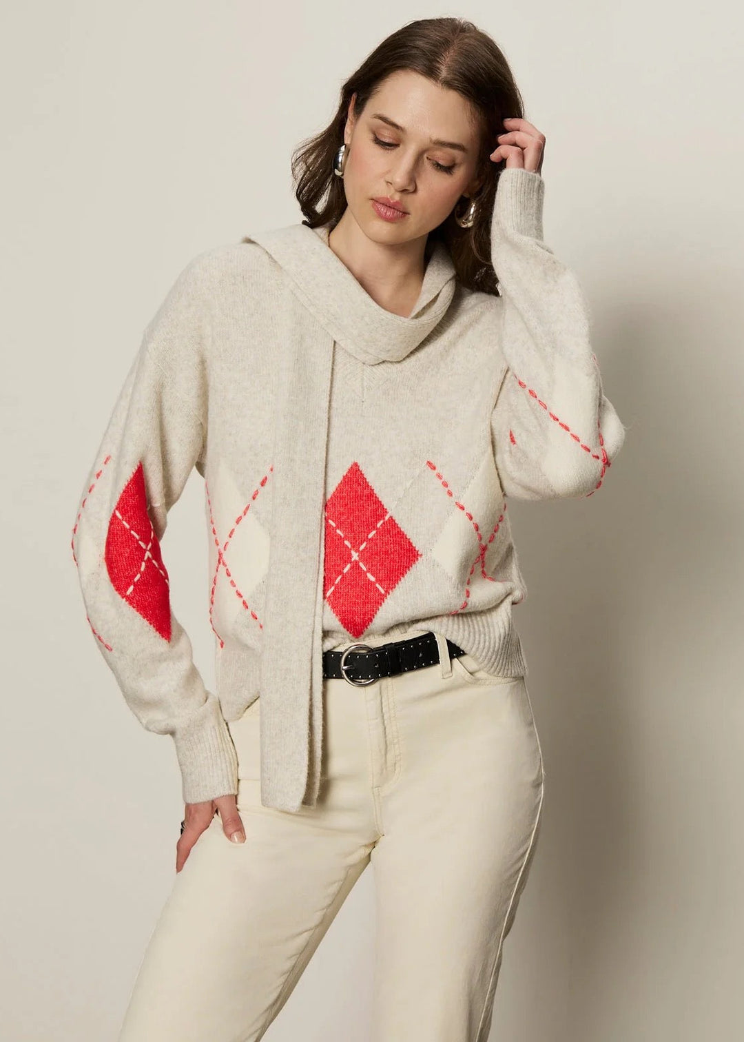 Woman wearing a cream sweater with red diamond patterns and white pants against a plain background