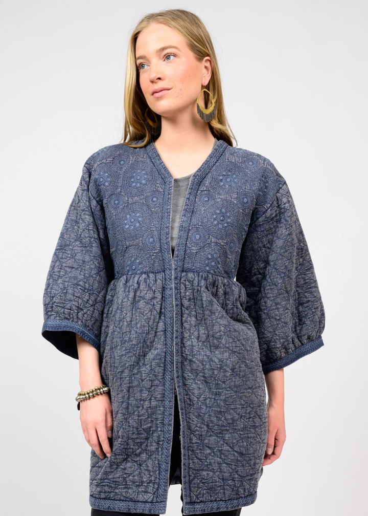 Woman wearing a long dark blue top with 3/4 length sleeves and a deep v-neck and floral embroidery standing in front of a white background