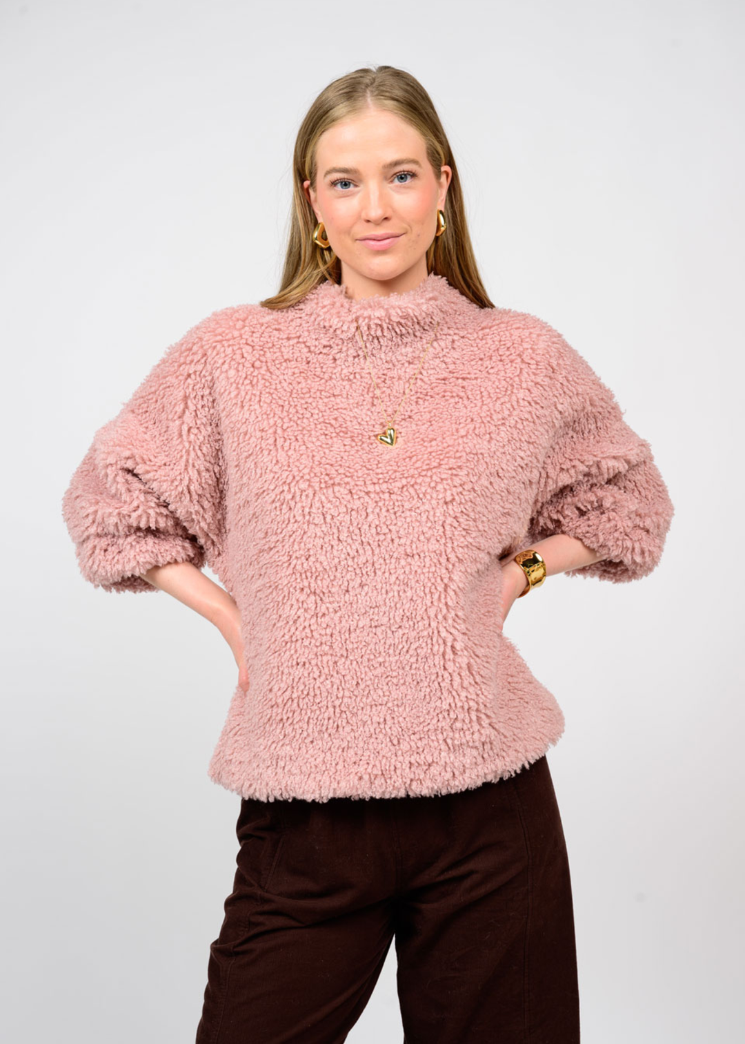 Woman with hands on hips wearing a pink sherpa top with 3/4 length sleeves and black jeans in front of a white background