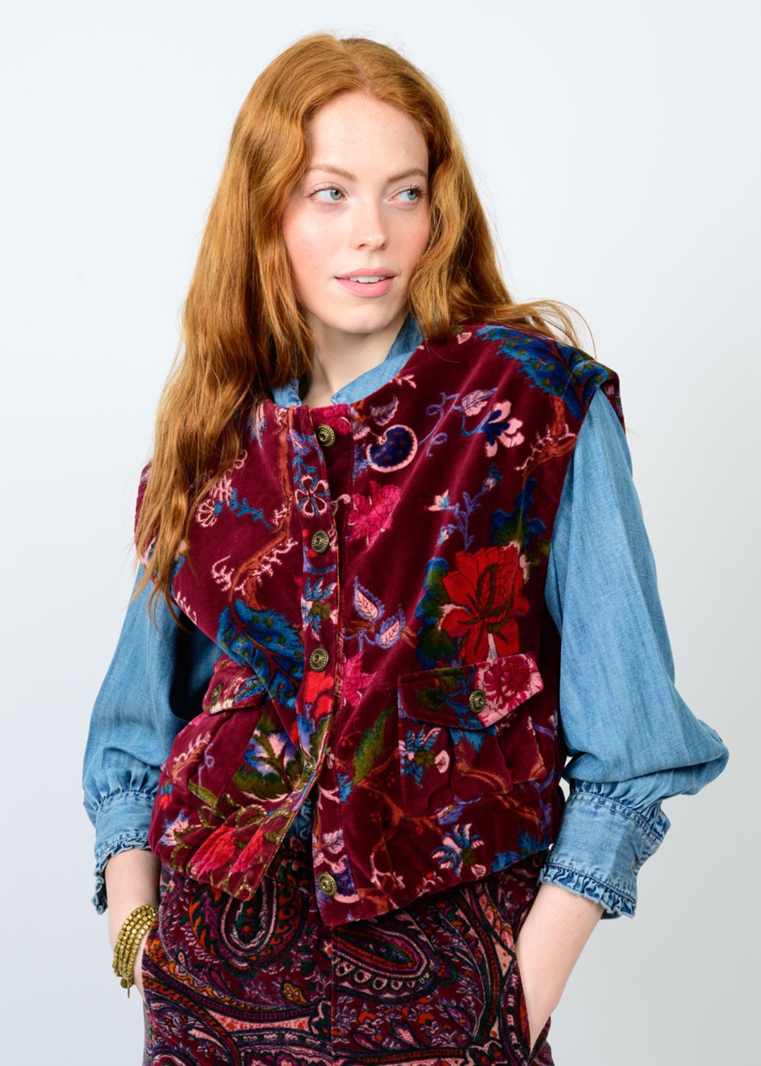 Woman with hands in pockets wearing a maroon floral vest against a white background