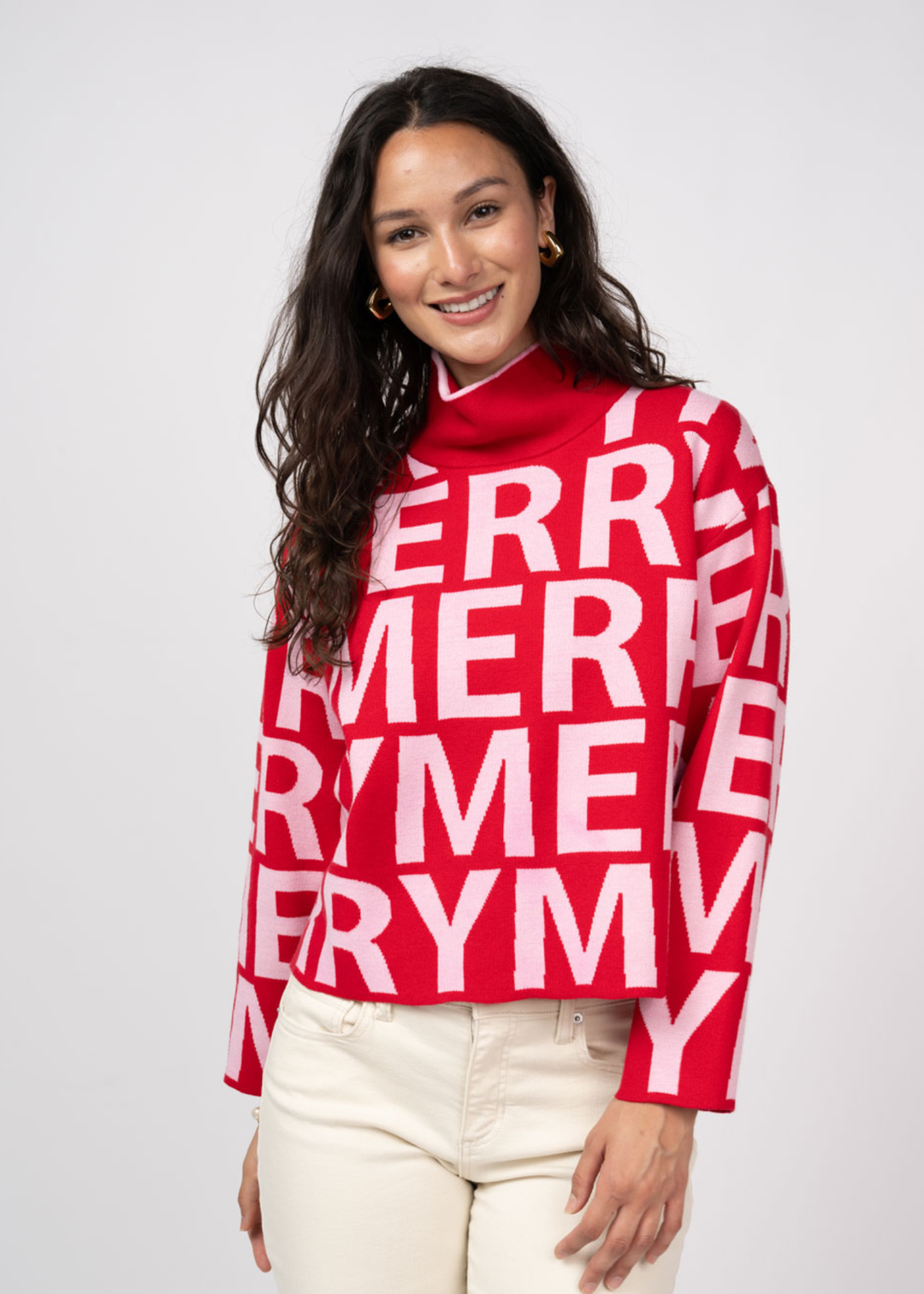 Woman wearing a red mock neck sweater with white "MERRY" letters and white jeans 
