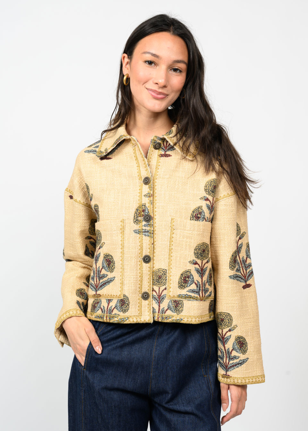 Woman with hand in pocket wearing a tan jacket with a floral print and navy pants in front of a white background