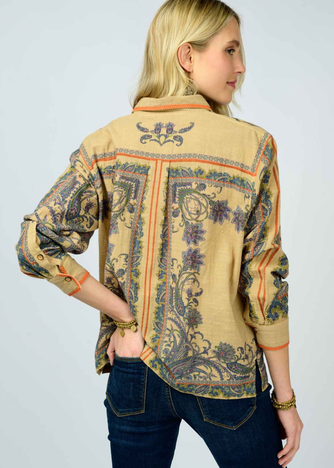 Woman turned around wearing a camel colored button down top with a floral print with dark denim jeans against a white background