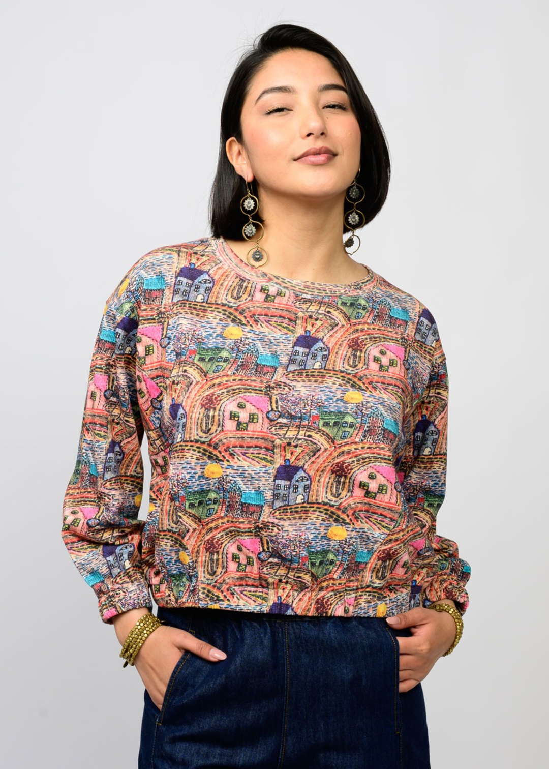 Woman wearing a colorful top with houses on it and dark pants against a white background