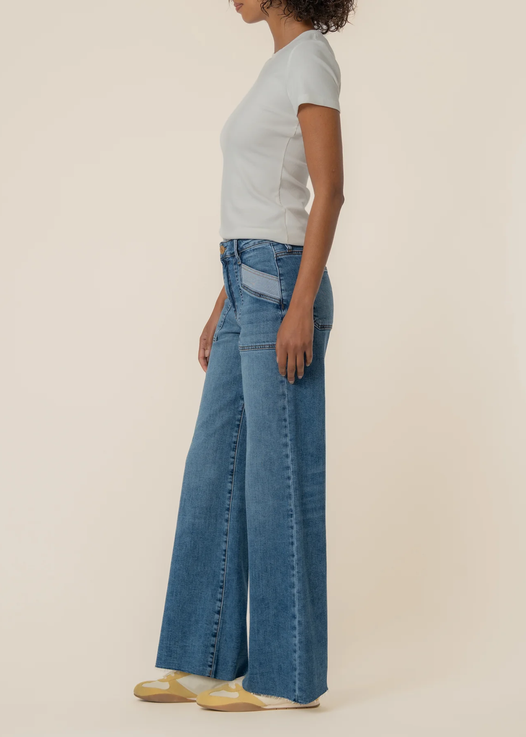 Woman turned to the side wearing blue wide leg jeans with a raw hem and front pockets against a tan background