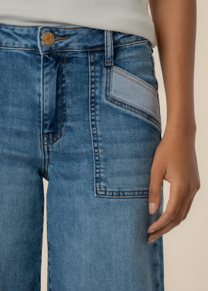 Close up of a woman wearing blue wide leg jeans with a raw hem and front pockets against a tan background