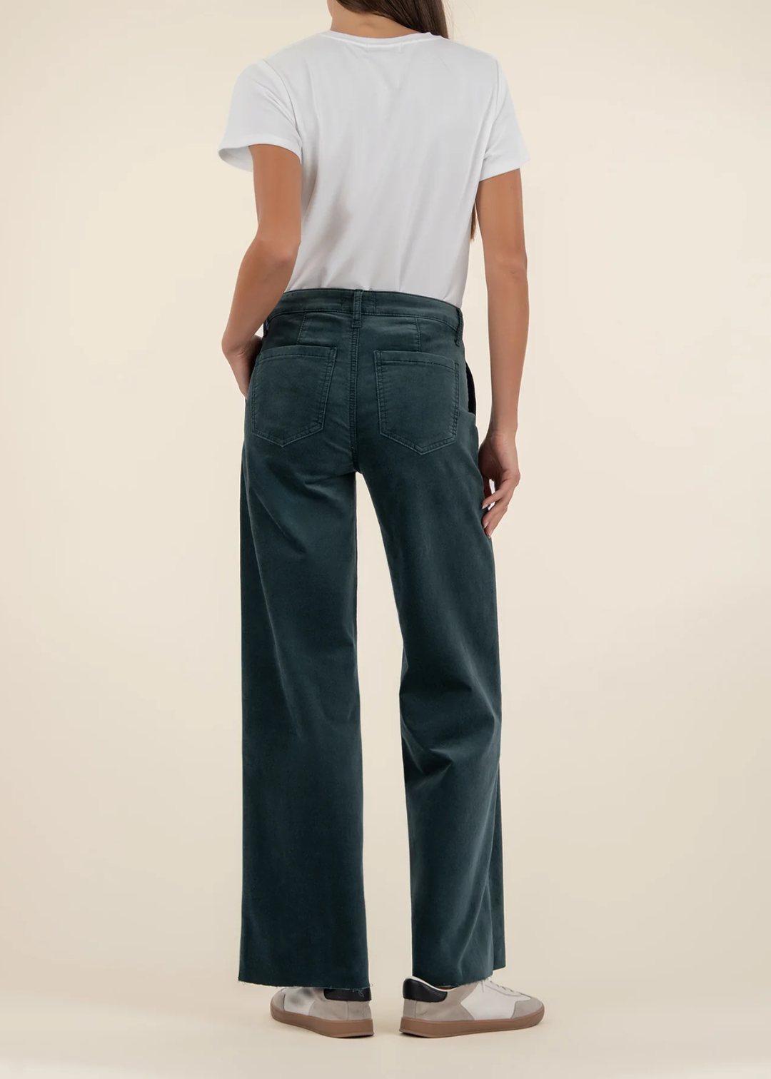 Woman turned around wearing teal velvet jeans with a white t-shirt and sneakers against a tan background