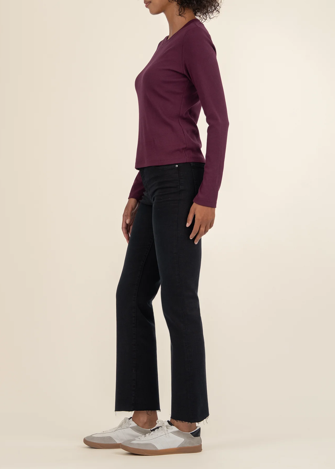 Woman turned to the side wearing a maroon long sleeve tee with black jeans and white sneakers in front of a white background