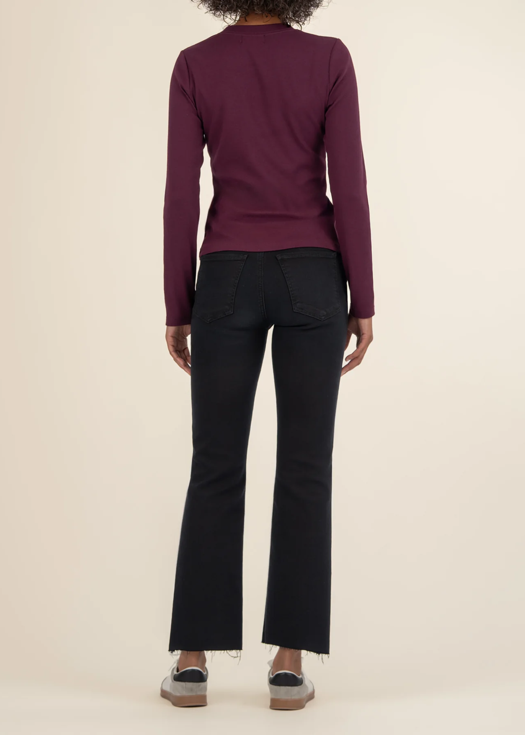Woman turned around wearing a maroon long sleeve tee with black jeans and white sneakers in front of a white background