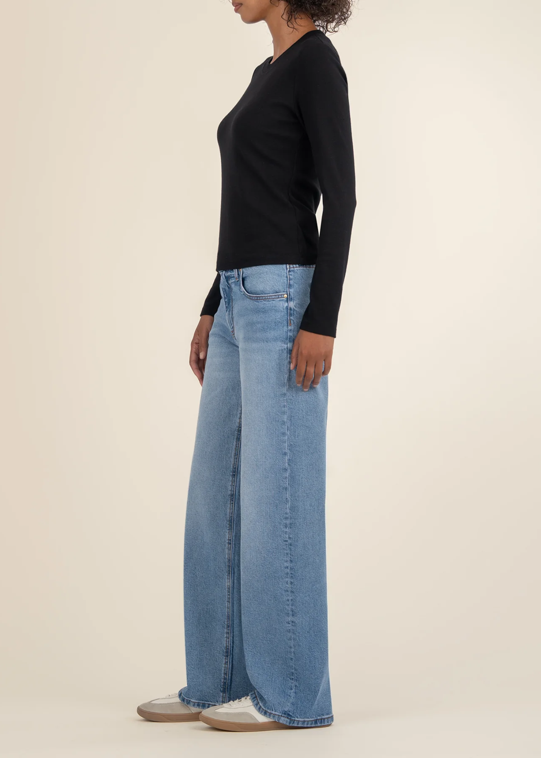 Woman turned to the side wearing a black long sleeve tee with jeans and white sneakers in front of a white background