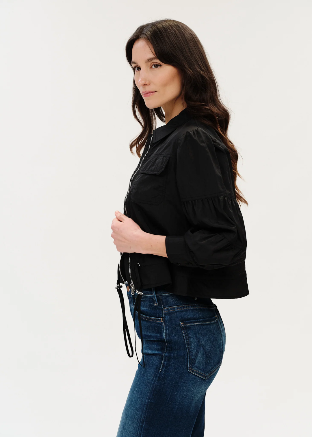 Woman with dark hair posing in black jacket with hand on the zipper turned to the side.