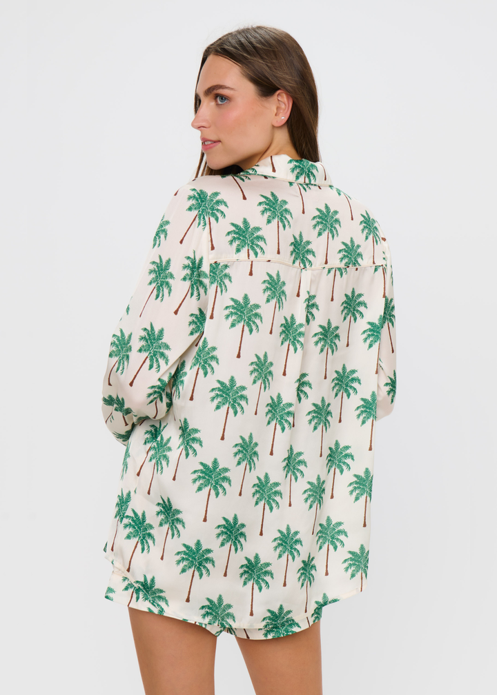 Woman turned around wearing a palm tree patterned pajama set with shorts and a long sleeve top against a white background