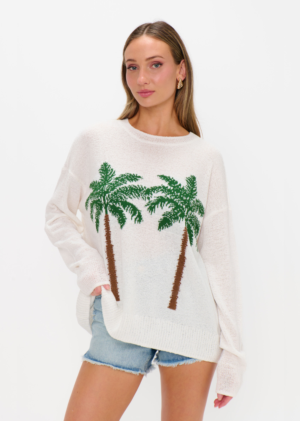 Woman wearing a white sweater with two palm trees on it and denim shorts against a white background