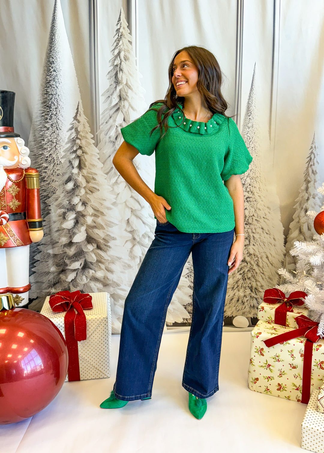 Woman in a green top and blue jeans standing in a festive setting with Christmas decorations.